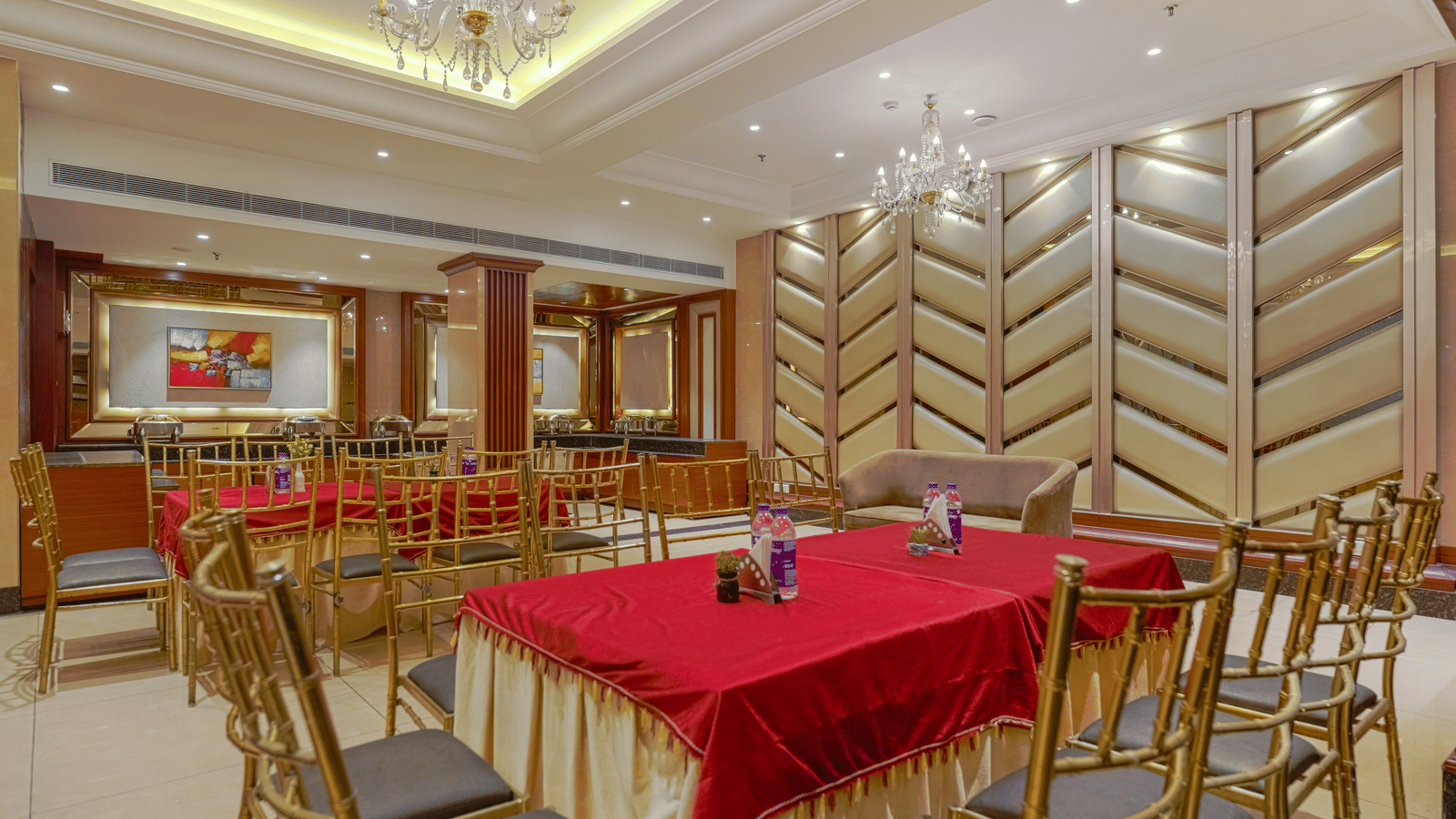 A dining room featuring tables with red cloths, gold chairs, and a wall with a textured, zig-zag padded design at BluSalzz Select City Centre, Amritsar.
