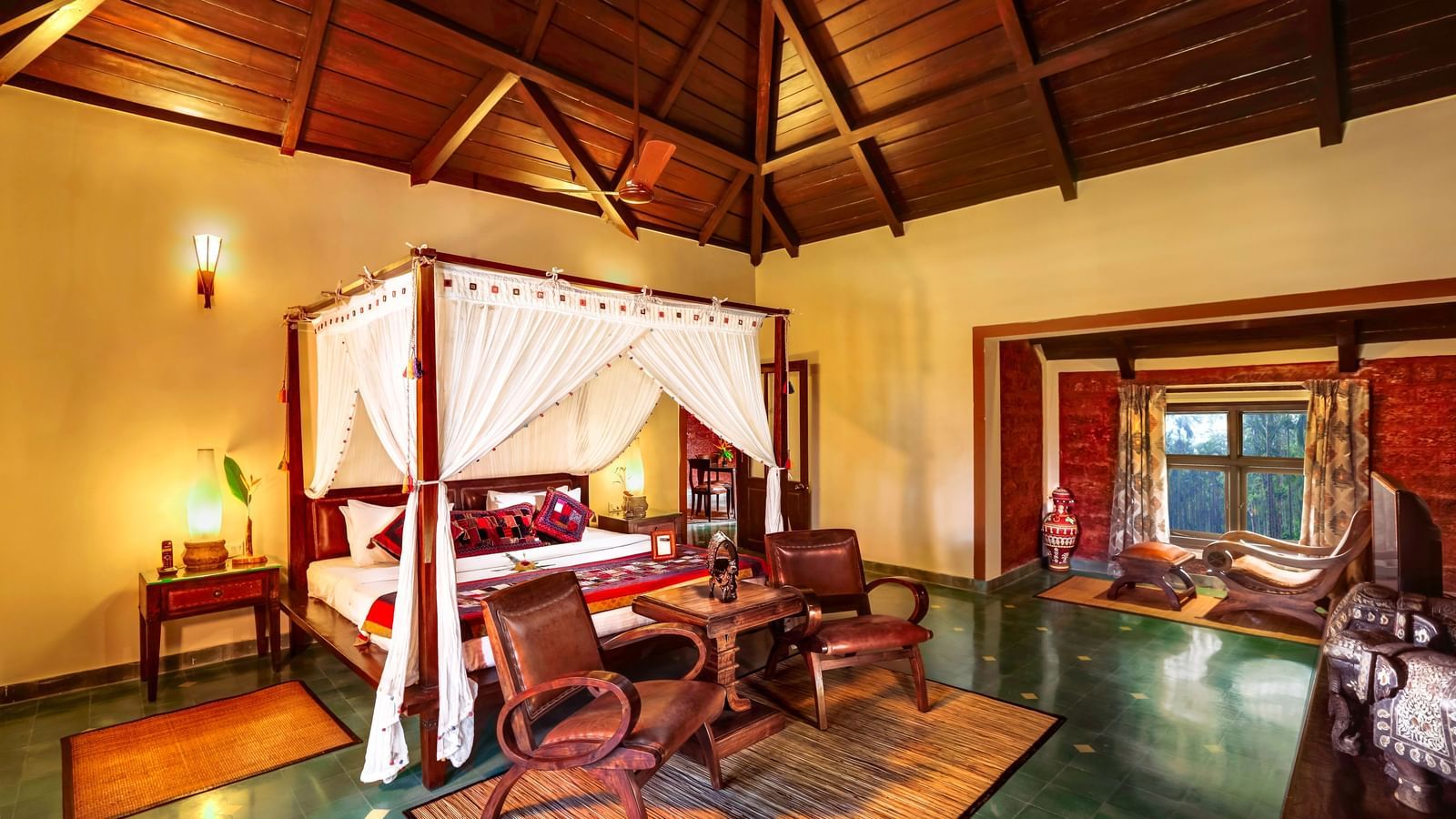 A warmly-lit room with a poster bed, sitting areas at its foot and a sitting space by the window at Hoysala Village Resort, Hassan