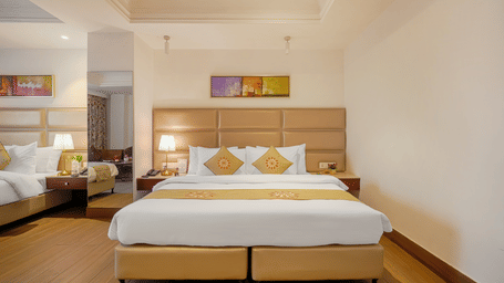 A view of an elegant hotel room featuring a large bed with a tan headboard, gold runner, and bright, white walls at BluSalzz Select City Centre, Amritsar.