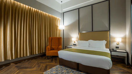 Polo Hotel, Agartala boutique room with king bed, orange wingback armchair, gold curtains, bedside lamps and patterned rug on wooden floor.