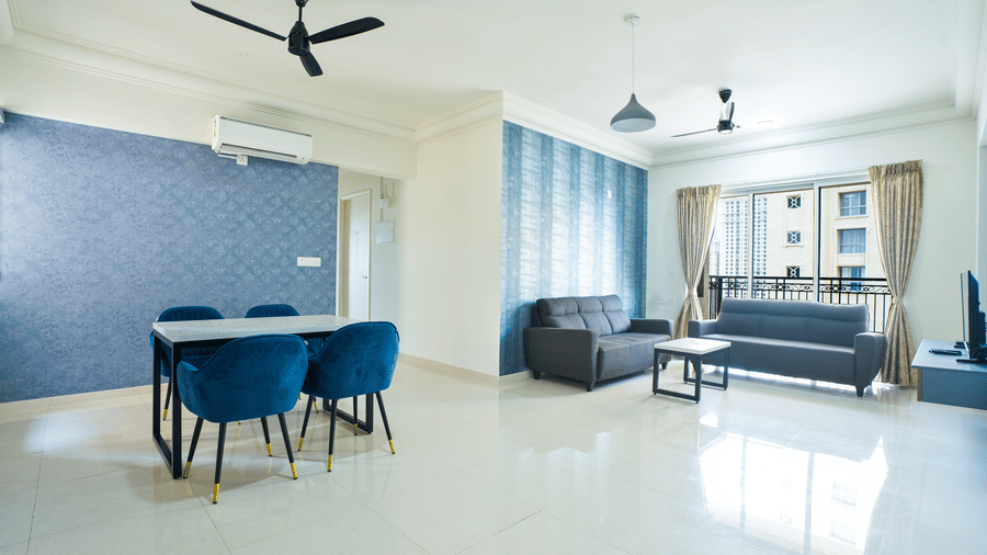 A bright, open-plan living and dining area with blue velvet chairs, a grey sofa, and a large window in a modern, tiled apartment at Apartel by Aarin, Oragadam.