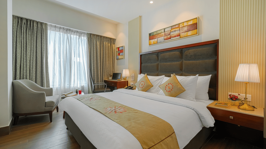A hotel room with a large bed, featuring golden textiles, modern art, and heavy curtains covering a wide window at BluSalzz Select City Centre, Amritsar.