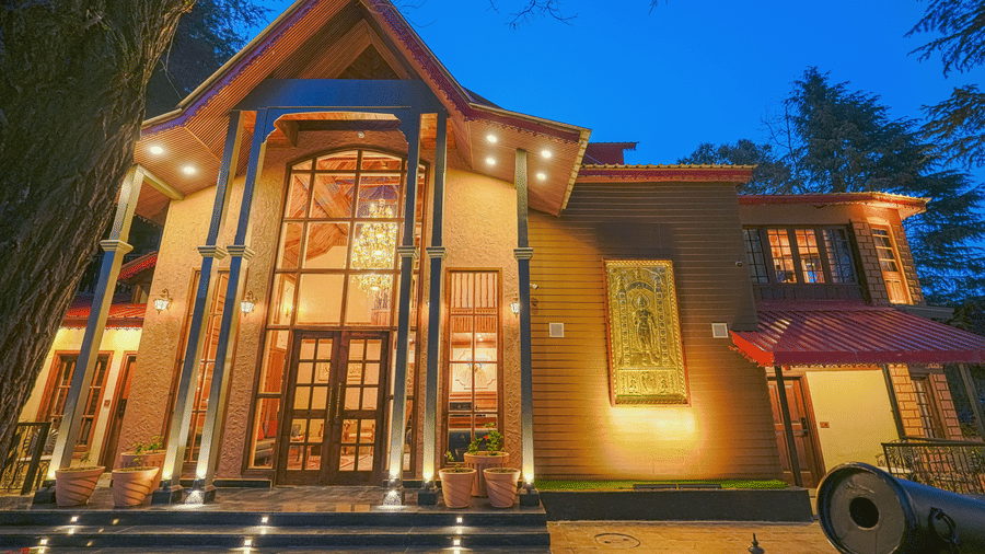 The well-lit facade of BluSalzz Terrah Hills Resort, Dalhousie, with steps in the entrance, a large wooden door, and trees in the backdrop at night.