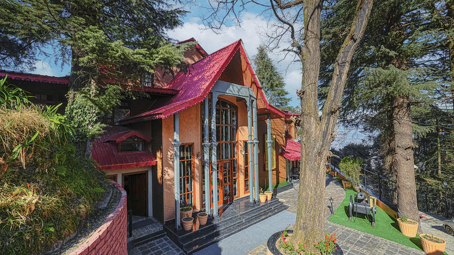 The facade of BluSalzz Terrah Hills Resort, Dalhousie, with a roof, surrounded by trees and a garden path.