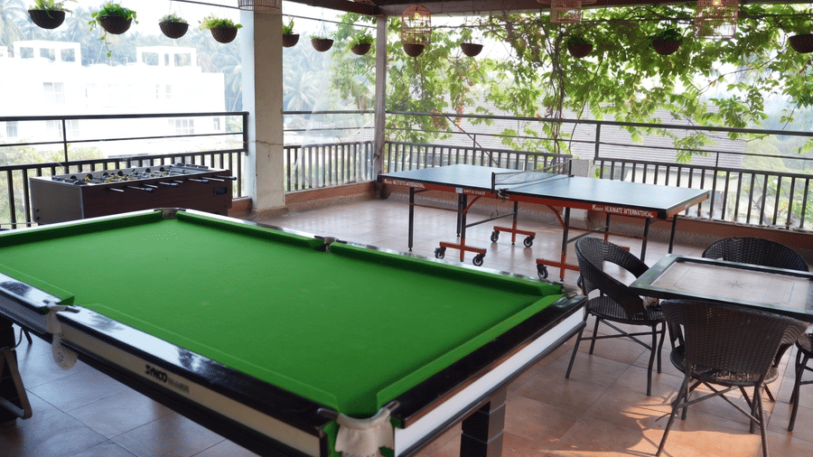 A pool table stands alongside a table tennis setup, seating arrangements, hanging planters on a covered terrace at Bougainvillea, Alibaug.