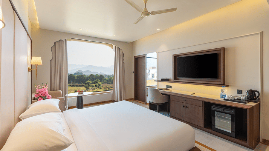 A room at EsthereaRaj Leela, Ranakpur, featuring a king size bed, a ceiling fan, a wall-mounted television, and a window overlooking green outdoor landscape view.