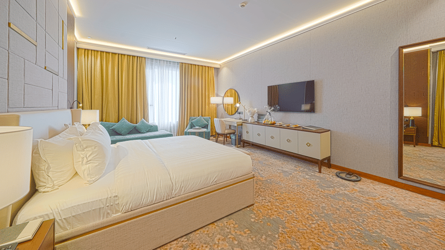 Deluxe King bedroom corner view showcasing the plush bed facing the wall-mounted TV, the work desk situated beside an upholstered couch in a corner | Grand Iva Hotel, Abha