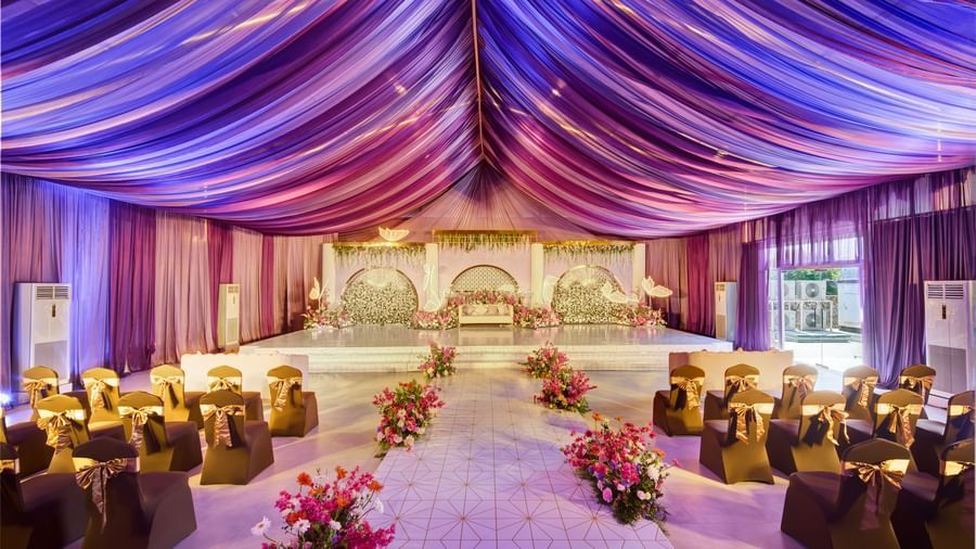 Decorated banquet hall with purple glowing roof, rows of chairs and flower centrepiece along the isle featuring a decorated stage at the backdrop.