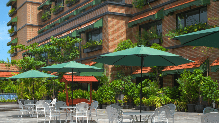 An outdoor seating area with green umbrellas, metal chairs and tables surrounded by plants and the building facade with balcony greenery at MAYFAIR Bay Resort, Paradeep.