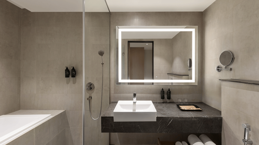 Luxurious hotel bathroom with a backlit vanity mirror, freestanding bathtub, sleek countertop basin, and grey stone-effect wall tiles.