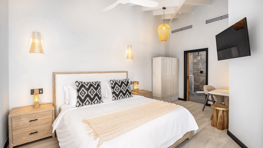 A bedroom at Tamarind Hills Resort and Villas, Antigua, with a king size bed with bedding, a lamp upon a nightstand, a cupboard, a wall mounted television and a door.