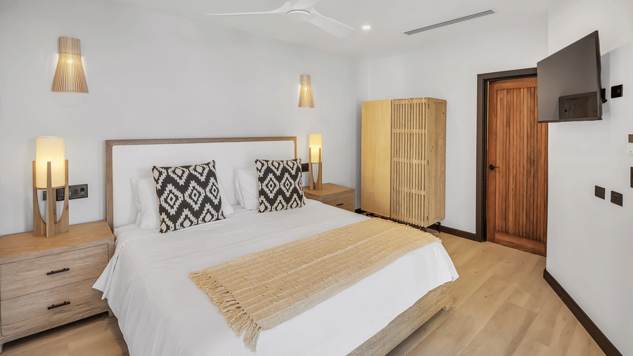 A bedroom at Tamarind Hills Resort and Villas, Antigua, with a king size bed with bedding, a lamp upon a nightstand, a cupboard, a wall mounted television and a door.