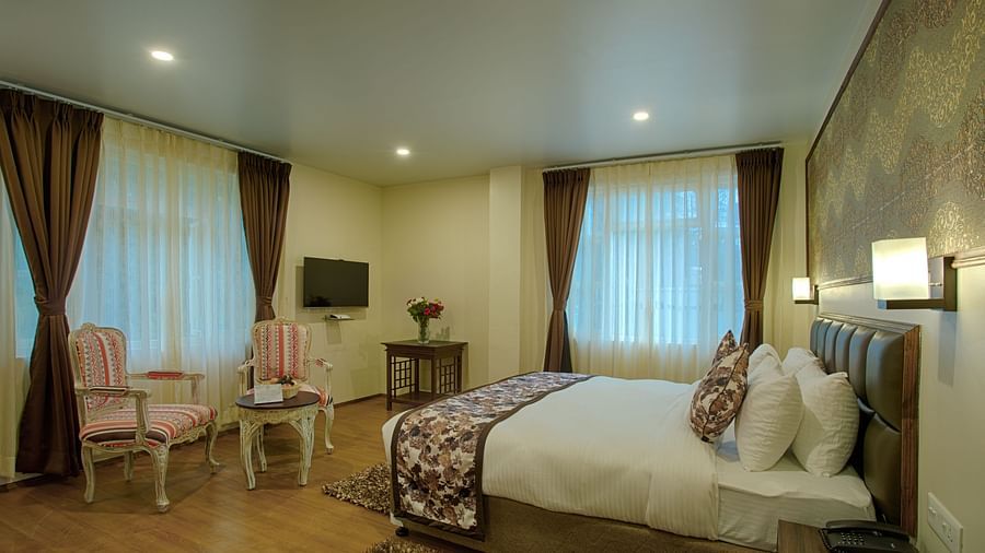 full view of suite room showcasing a king size bed, 2 chairs and a teapoy, a television and much more along with curtained windows - the aryan regency