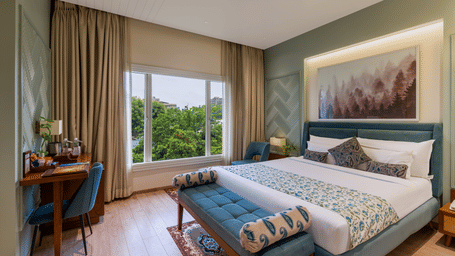 Full view of a brightly lit Premium Room with a teal-accented bed, a wooden workstation by the window, and polished wooden floors - Amantra Comfort Hotel