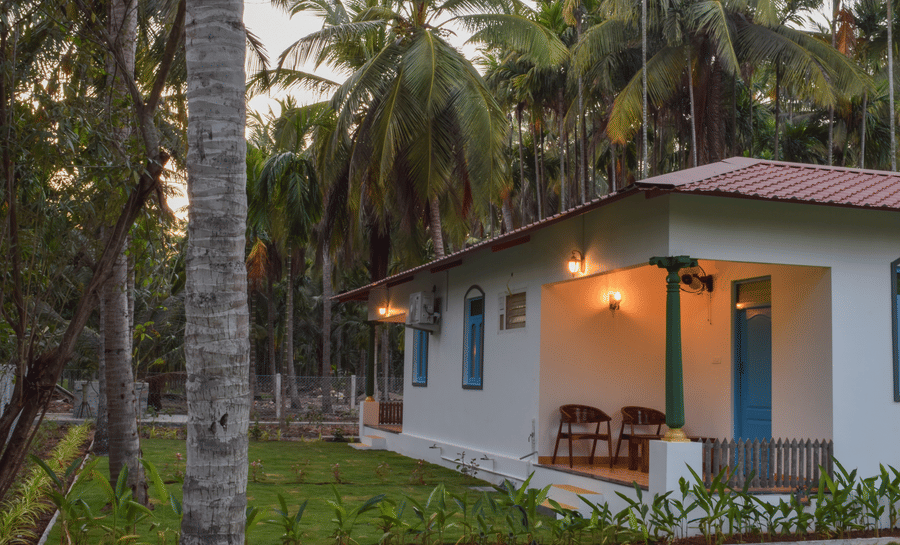 A white resort cottage with a red roof and blue window frames, surrounded by palm trees and green lawns - Ibex Resorts, Coimbatore (Kakarla)