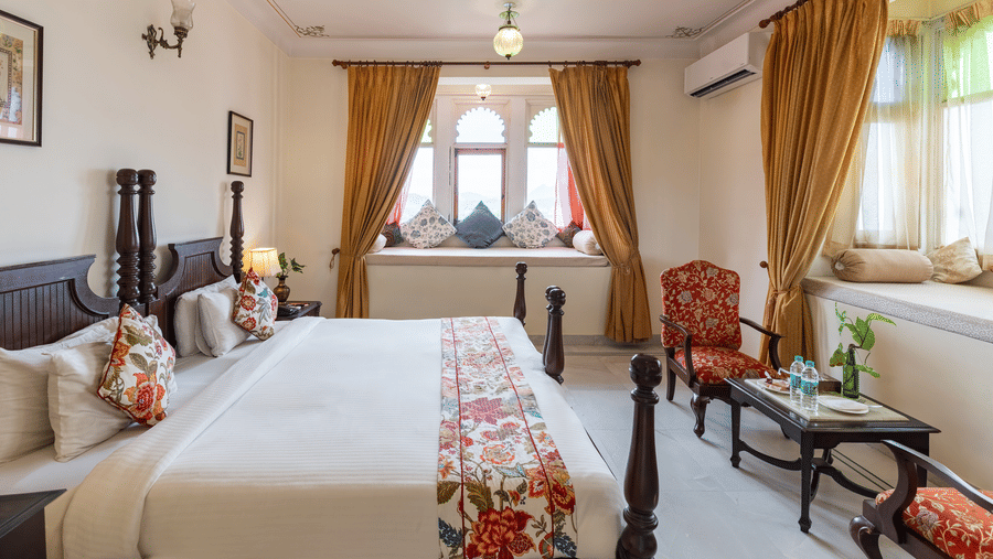 A bedroom with twin beds, curtained windows, seating area with 2 chairs and table, and patterned armchairs at Akshay Niwas Boutique Hotel By Amantra, Udaipur.