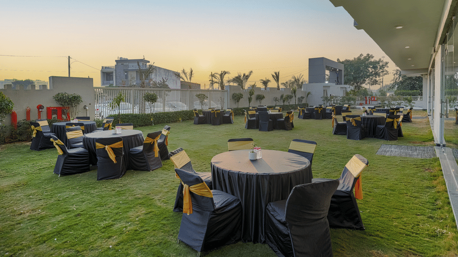 A rooftop venue at Urban Suites by BluSalzz, Jaipur, with a grassy area, set tables with chairs, and a city skyline in the background at dusk.