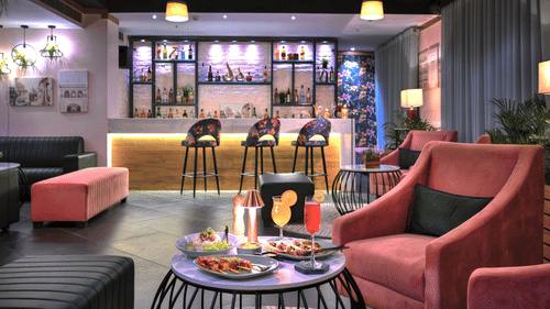 An image with a coffee table with food in the foreground and a bar counter with high chairs in the background  - Rooftop in Agra - Golden Tulip, Agra