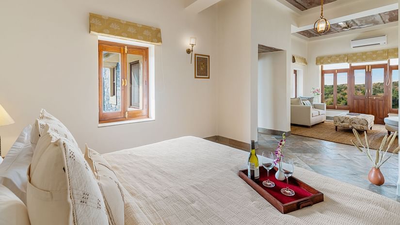 A king bed with a tray of that consists of a bottle of bubbly drink and champagne glasses on the bed inside one of the rooms - two chairs and a small table in between them on a balcony inside a room - Chunda Shikar Oudi, Udaipur