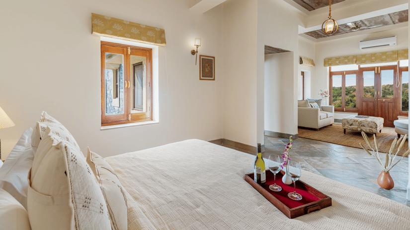A king bed with a tray of that consists of a bottle of bubbly drink and champagne glasses on the bed inside one of the rooms - two chairs and a small table in between them on a balcony inside a room - Chunda Shikar Oudi, Udaipur