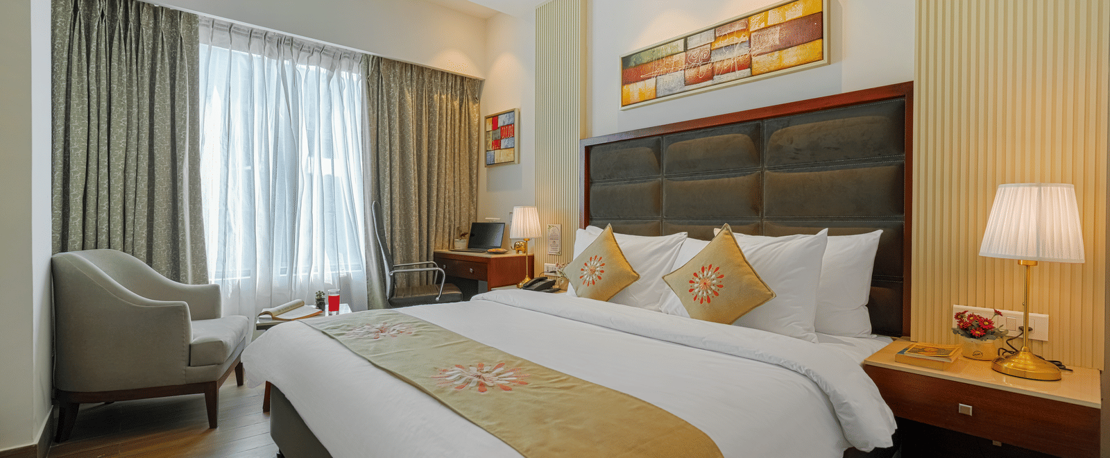 A hotel room with a large bed, featuring golden textiles, modern art, and heavy curtains covering a wide window at BluSalzz Select City Centre, Amritsar.