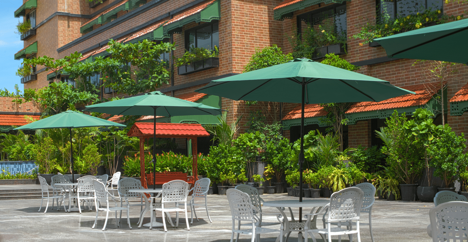 An outdoor seating area with green umbrellas, metal chairs and tables surrounded by plants and the building facade with balcony greenery at MAYFAIR Bay Resort, Paradeep.