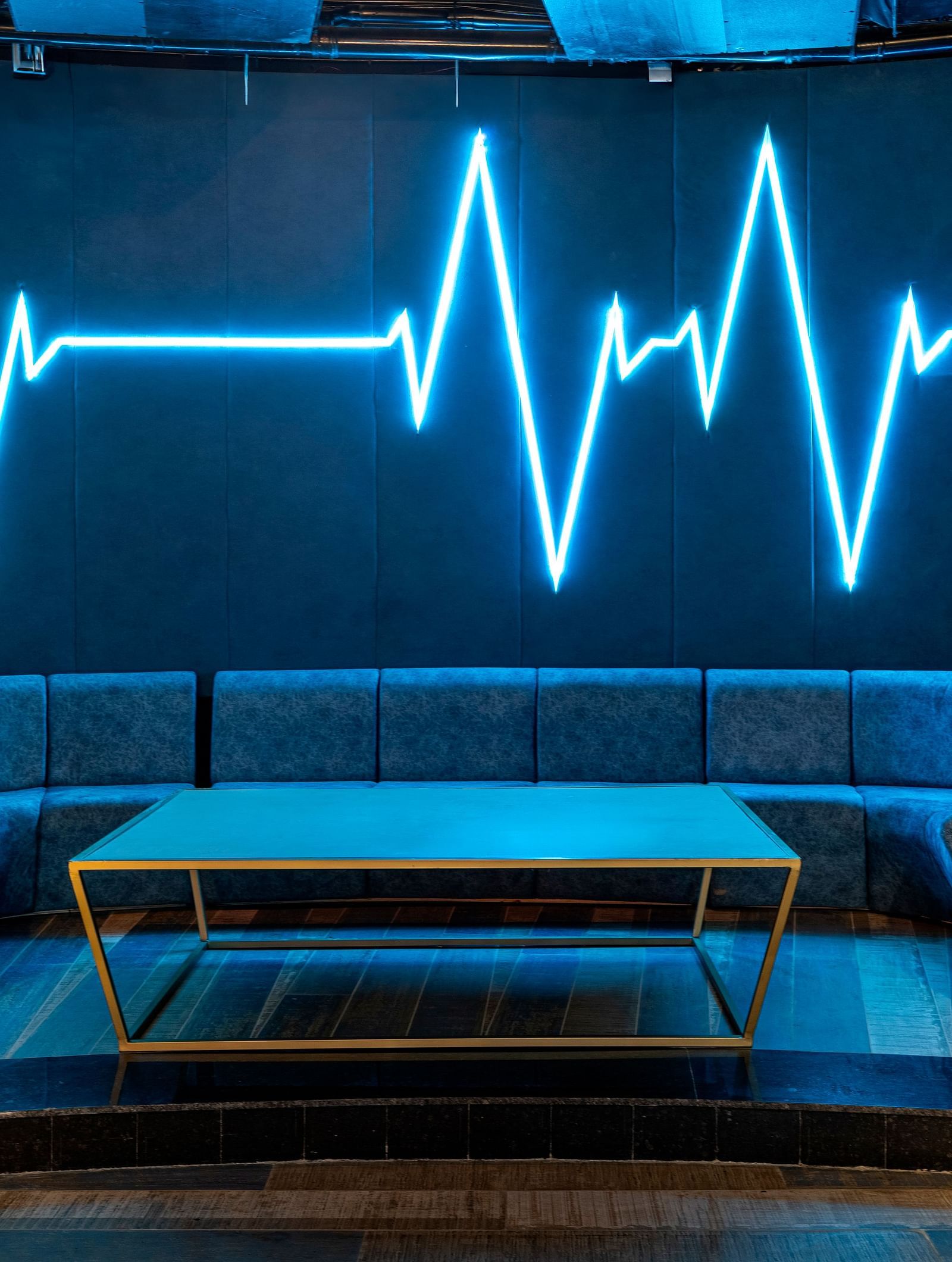 Verve with blue neon light in the shape of a heartbeat line on the wall, with curved seating and a small table at The Qualia Resort Club & Brewery, Udaipur.