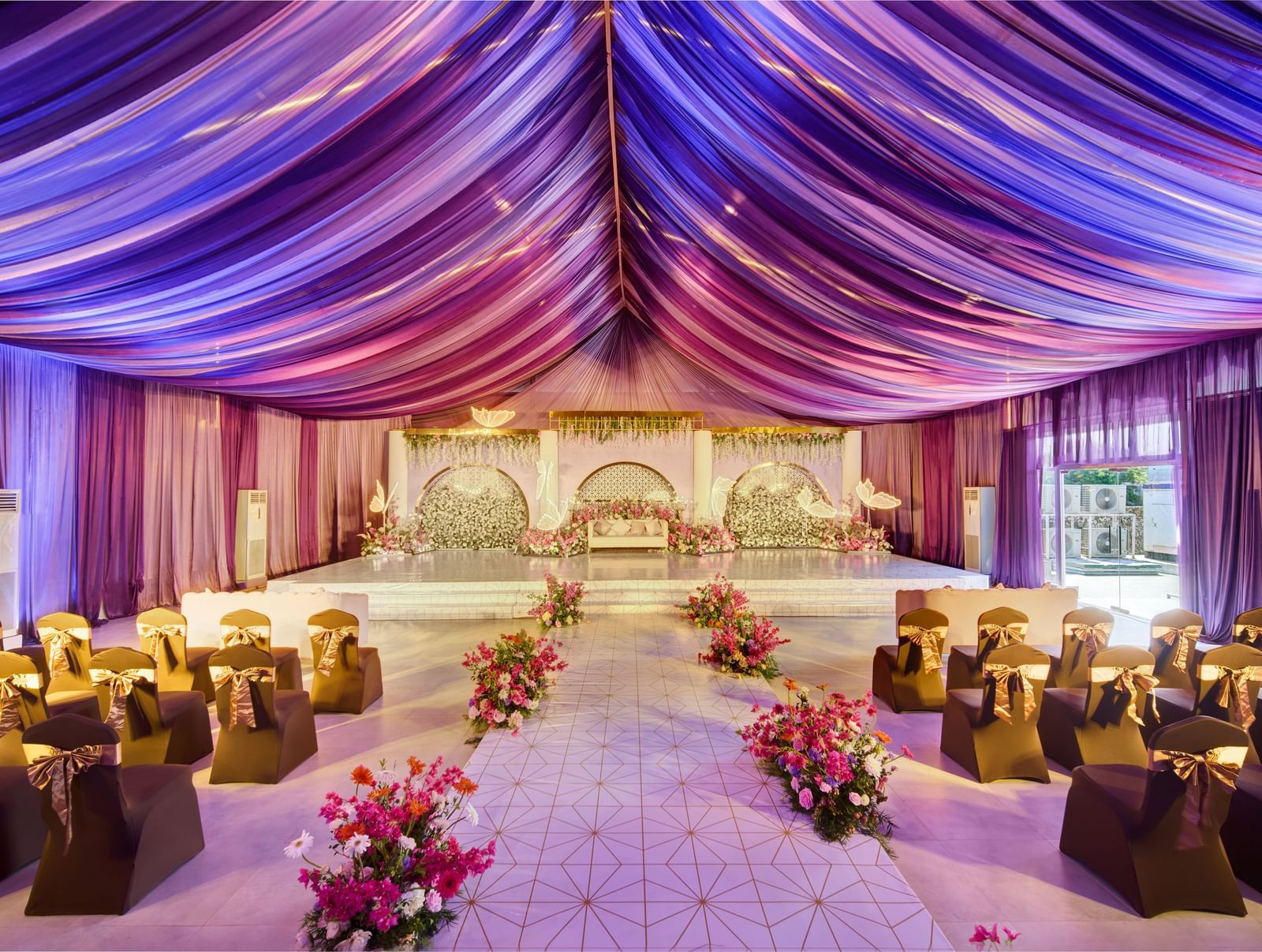 Decorated banquet hall with purple glowing roof, rows of chairs and flower centrepiece along the isle featuring a decorated stage at the backdrop.