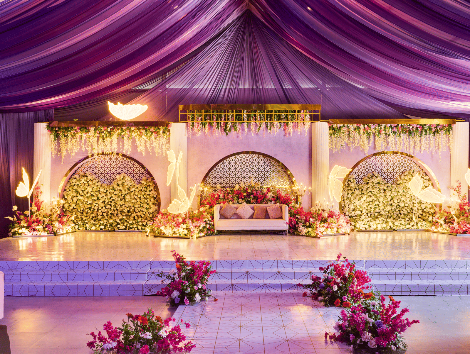 Floral wedding stage featuring a purple draped ceiling, a large chair for the bride and groom, and illuminated decorative arches - Grande Bay Resort & Spa, Mamallapuram