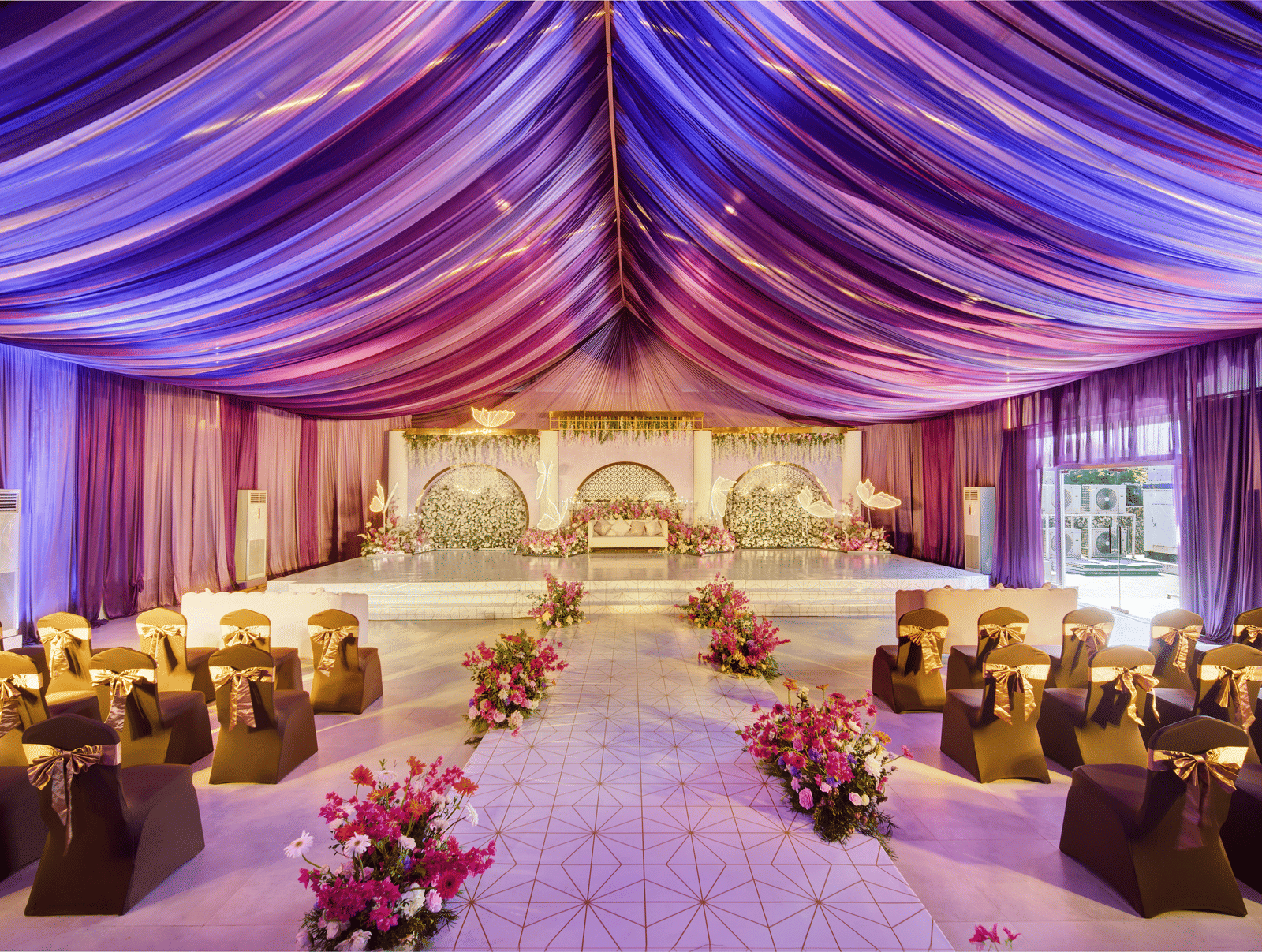 An indoor event venue in Mahabalipuram with a centred aisle framed by purple drapes leading to a floral wedding stage - Grande Bay Resort & Spa, Mamallapuram