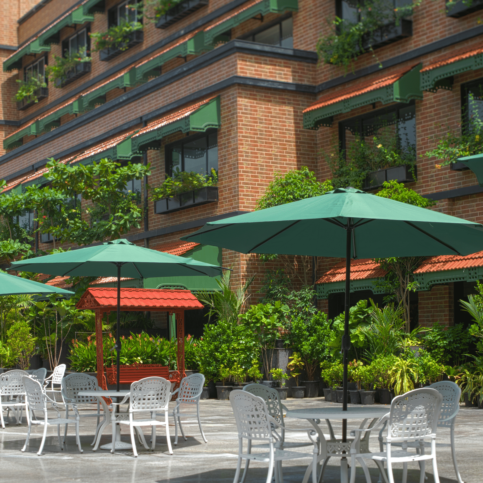 An outdoor seating area with green umbrellas, metal chairs and tables surrounded by plants and the building facade with balcony greenery at MAYFAIR Bay Resort, Paradeep.