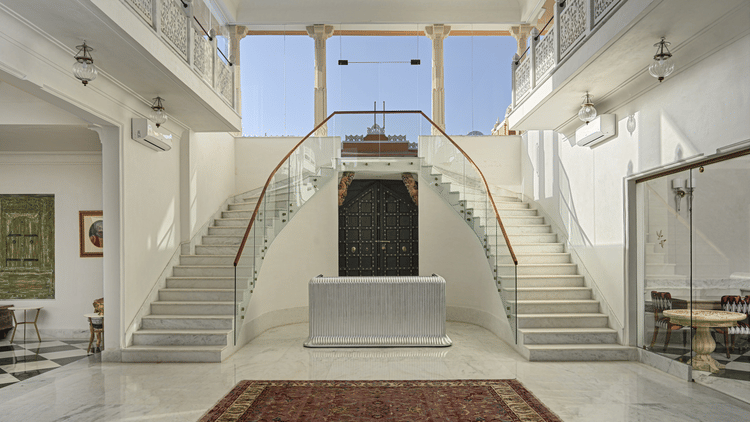 Interior view of a reception at Khas Mahal Suites, Udaipur featuring a symmetrical, double staircase with white risers and stringers, glass railings, a dark central door, a brown rug on the floor, and natural light entering from a high window.