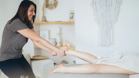 A smiling female masseuse in a gray shirt and dark pants is performing a leg massage on a client lying face down on a table.