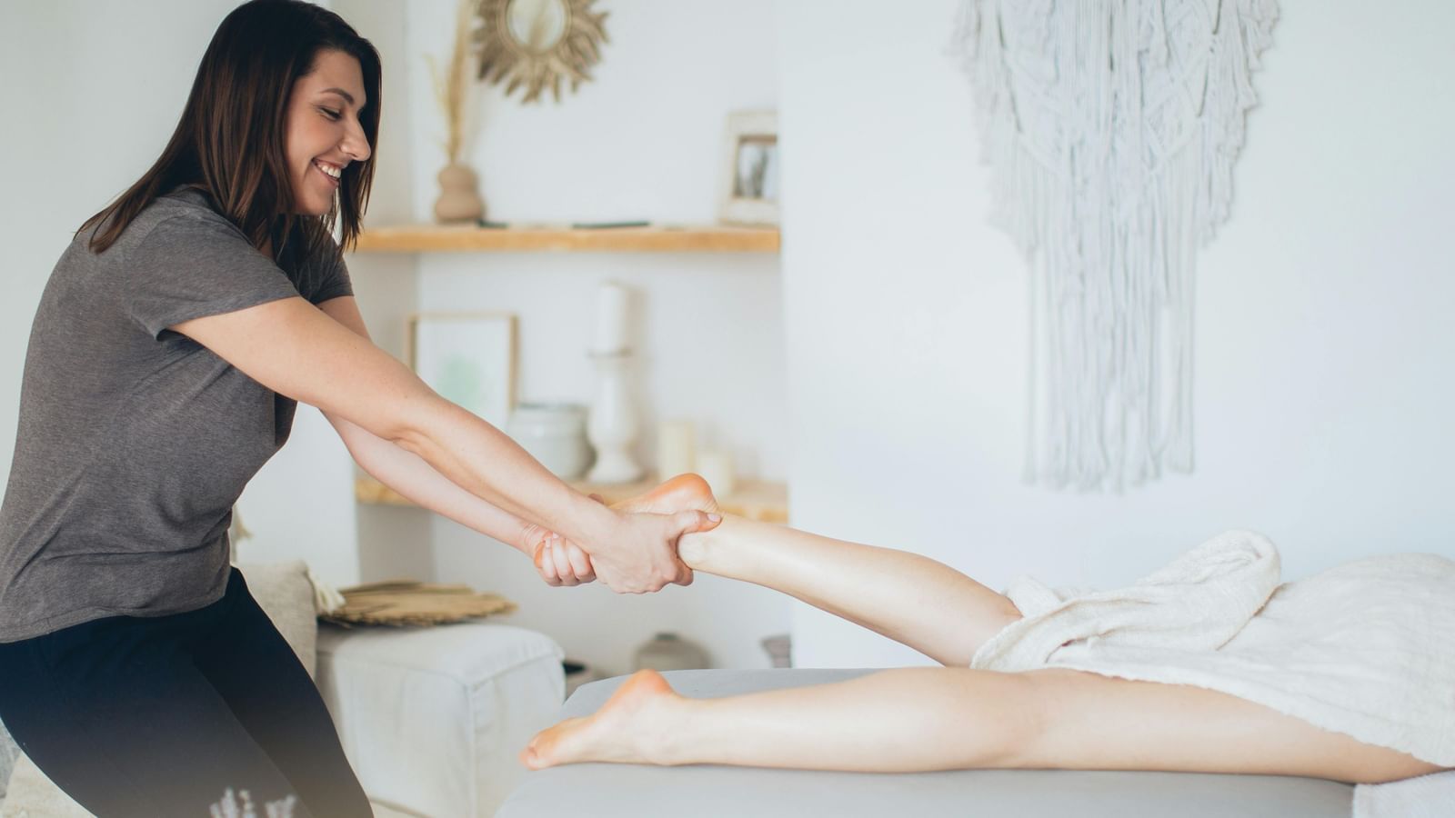 A smiling female masseuse in a gray shirt and dark pants is performing a leg massage on a client lying face down on a table.