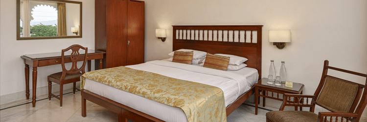 A spacious guest room at Ram Pratap Palace with a large bed, two chairs, a wardrobe, a desk, a mirror, and two bedside tables with lamps.