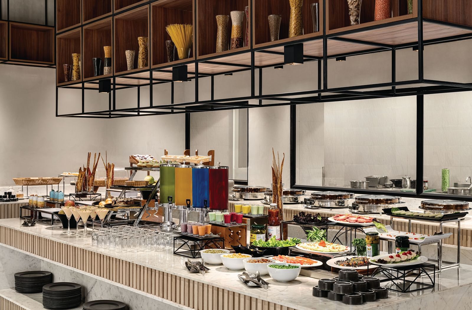 Ramada Encore by Wyndham, Bhiwadi | Buffet table with colorful food. Decorative metal structure above, high ceilings, large windows.