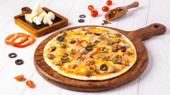 A round pizza topped with meat, olives, and vegetables is on a wooden board, surrounded by fresh tomatoes, cheese wedges, and spices.