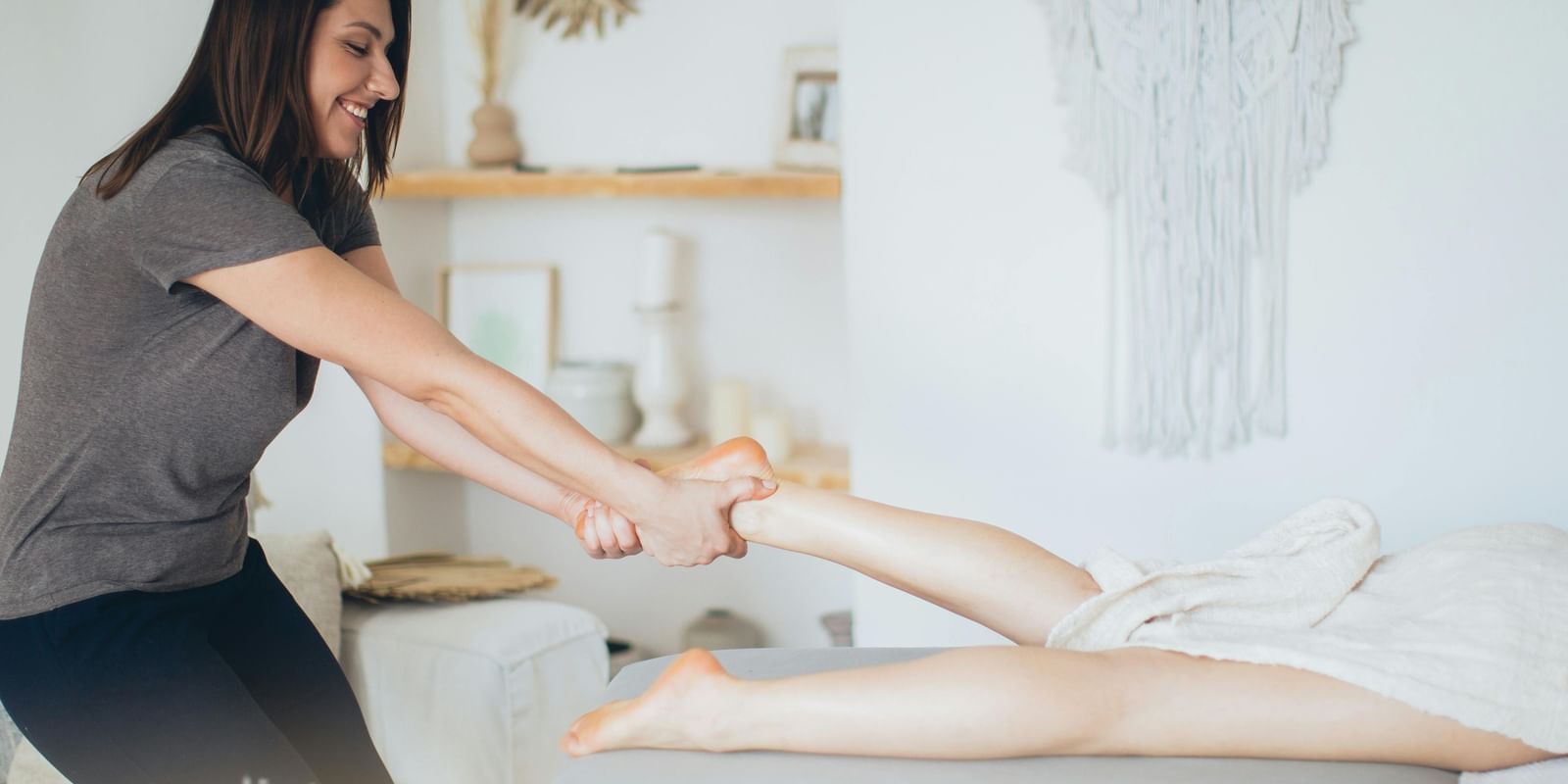 A smiling female masseuse in a gray shirt and dark pants is performing a leg massage on a client lying face down on a table.