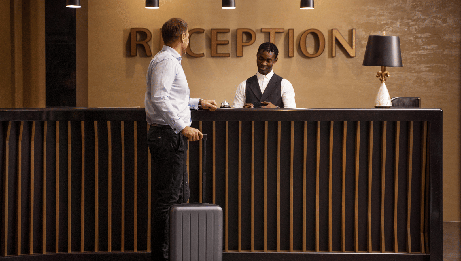 A guest with luggage stands at the hotel reception desk while a staff assists him.