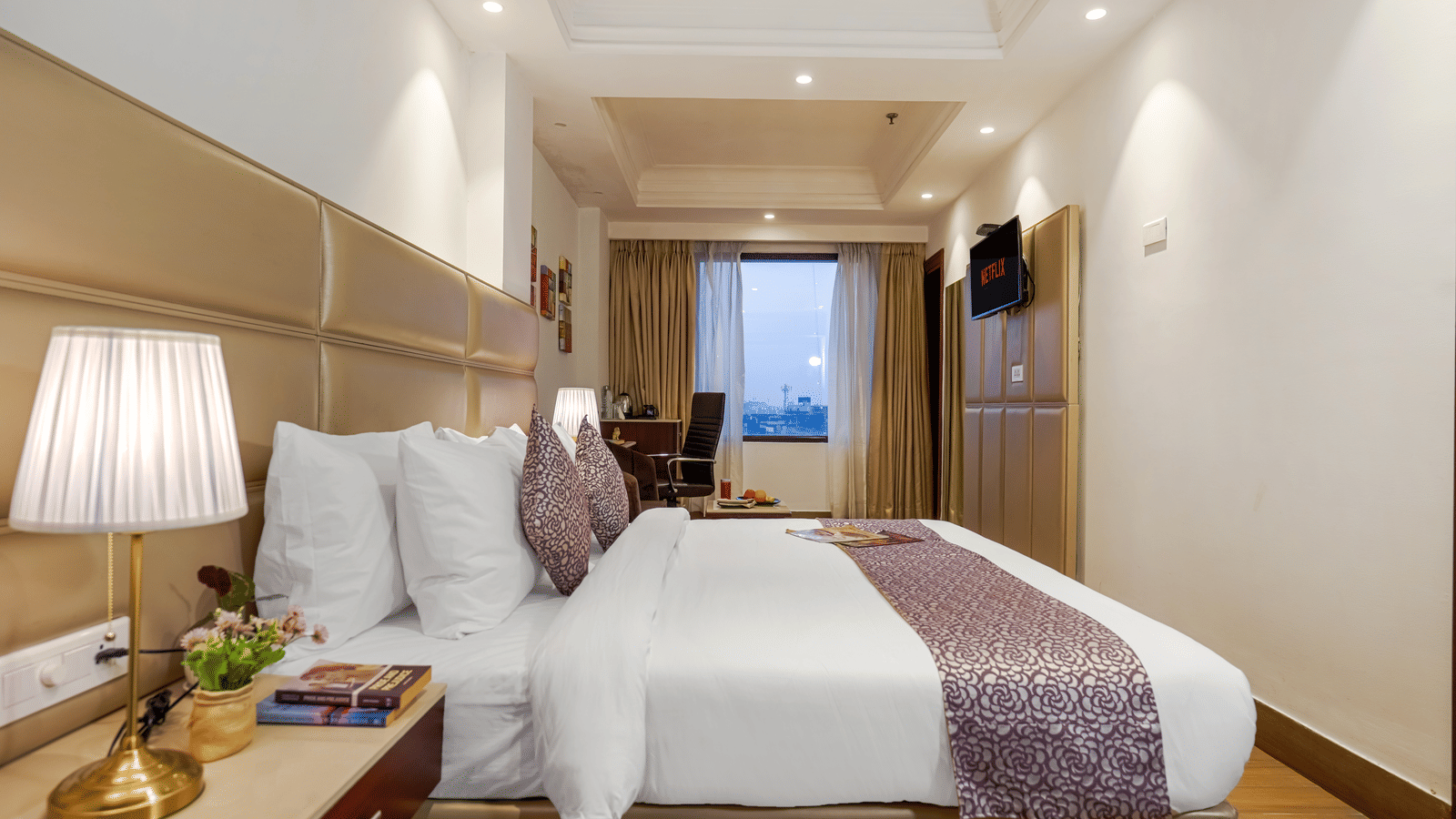 A modern, elegant hotel bedroom featuring a large bed with a patterned purple runner and a view of the city outside at BluSalzz Select City Centre, Amritsar.