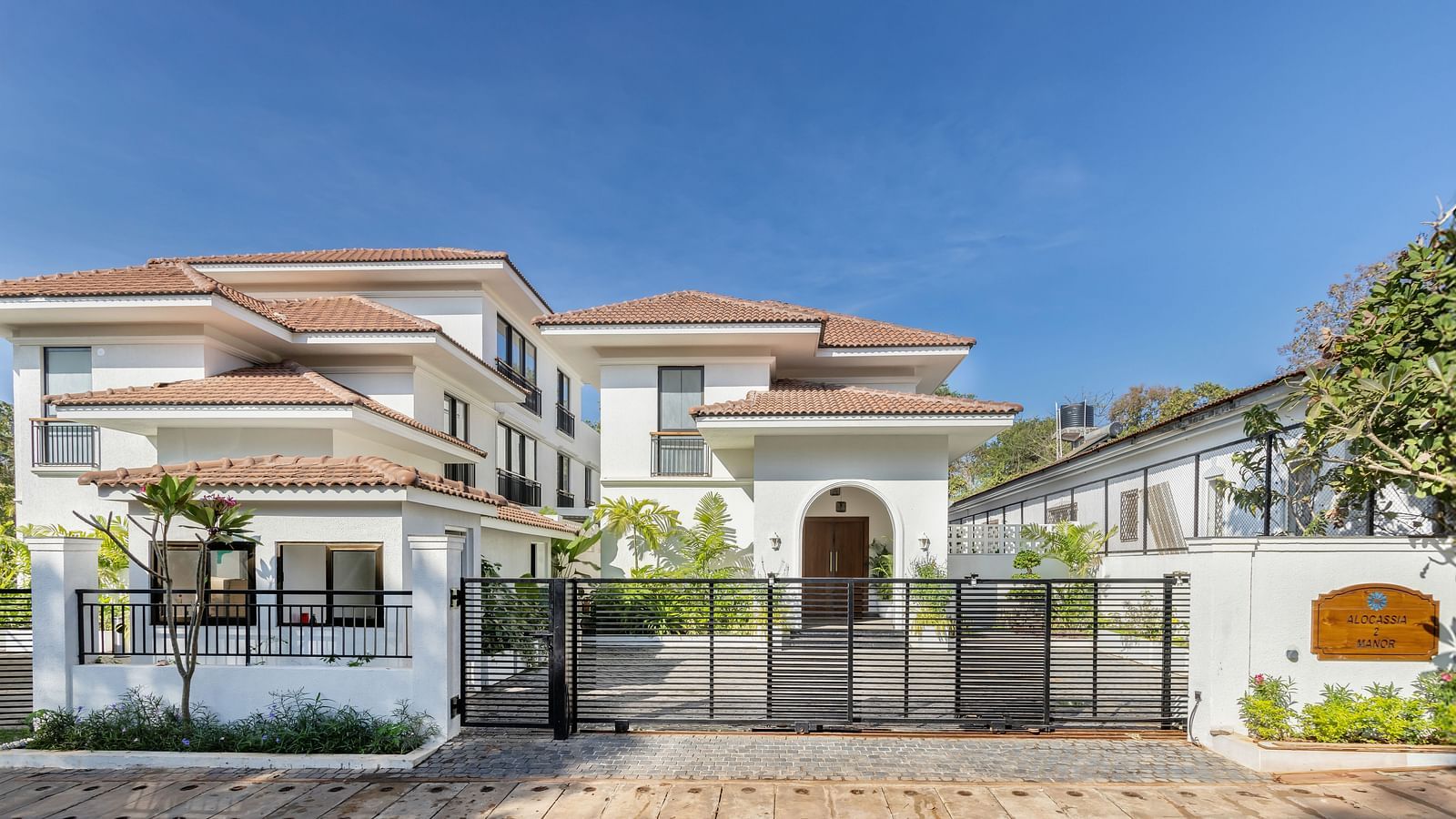 A wide-angle view of a luxurious villa with a spacious driveway, surrounded by well-maintained gardens and bright blue skies - BluSalzz Villas - Alocassia, Anjuna