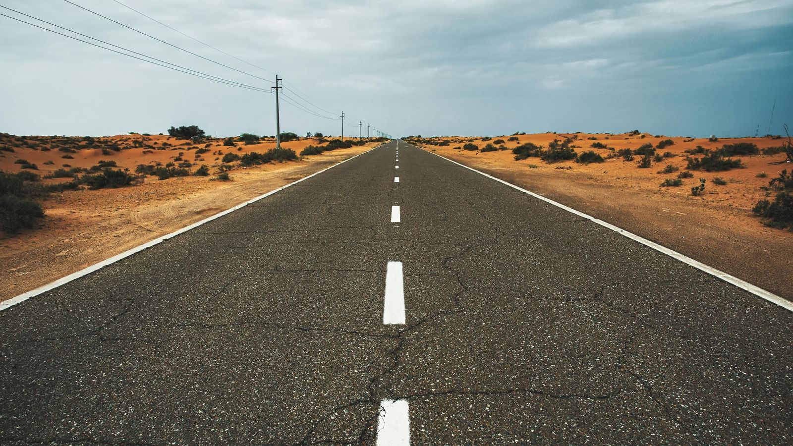 A long, straight road stretches to the horizon through a dry, desert landscape with scrub and telephone poles under a cloudy, dramatic sky.