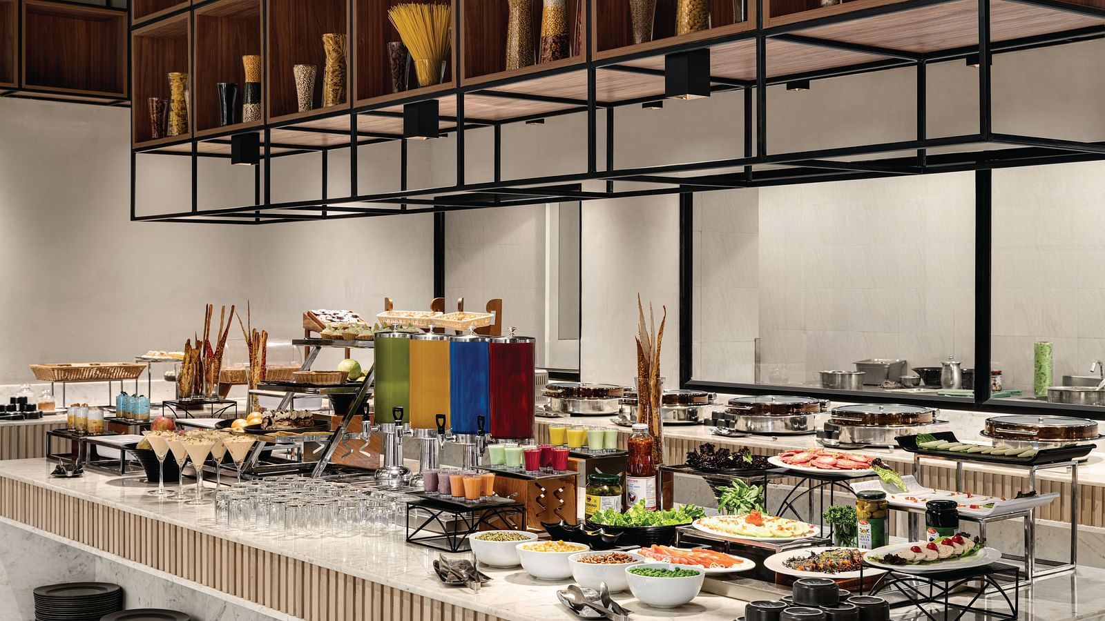 Ramada Encore by Wyndham, Bhiwadi | Buffet table with colorful food. Decorative metal structure above, high ceilings, large windows.