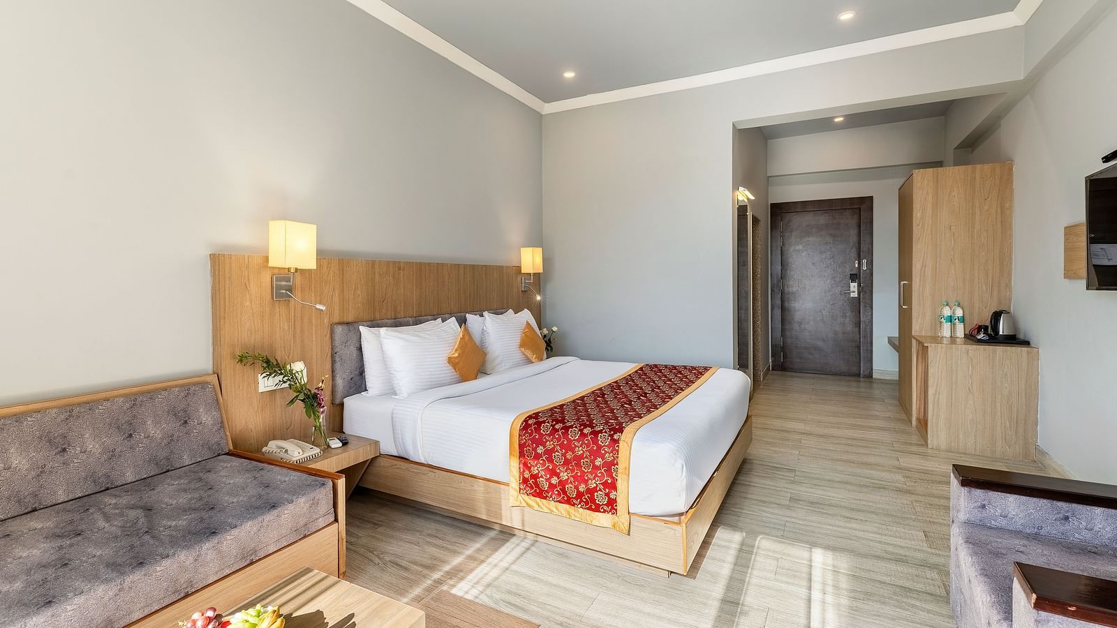 A tidy hotel room featuring a neatly made bed with a red runner, a gray sofa and a wooden side table with a fruit bowl on top of it - Via Lakhela Resort & Spa, Kumbhalgarh