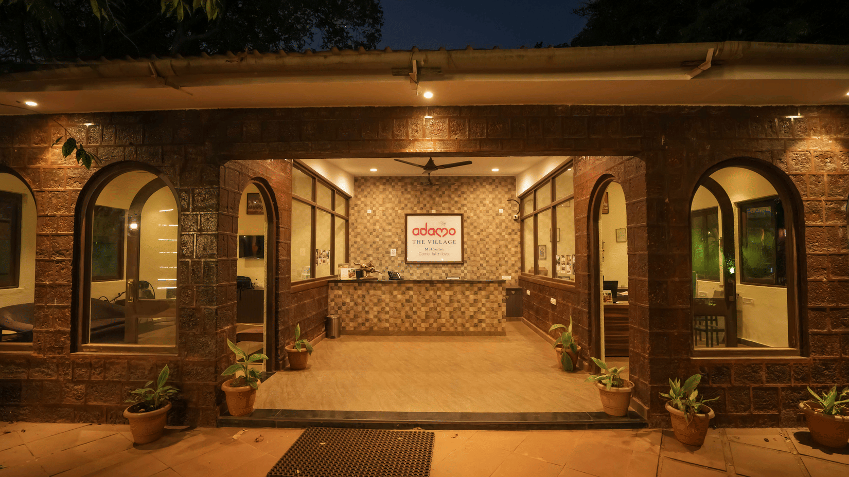 Night view of the Adamo The Village reception area exterior with a prominent front desk and arched openings set into stone walls.