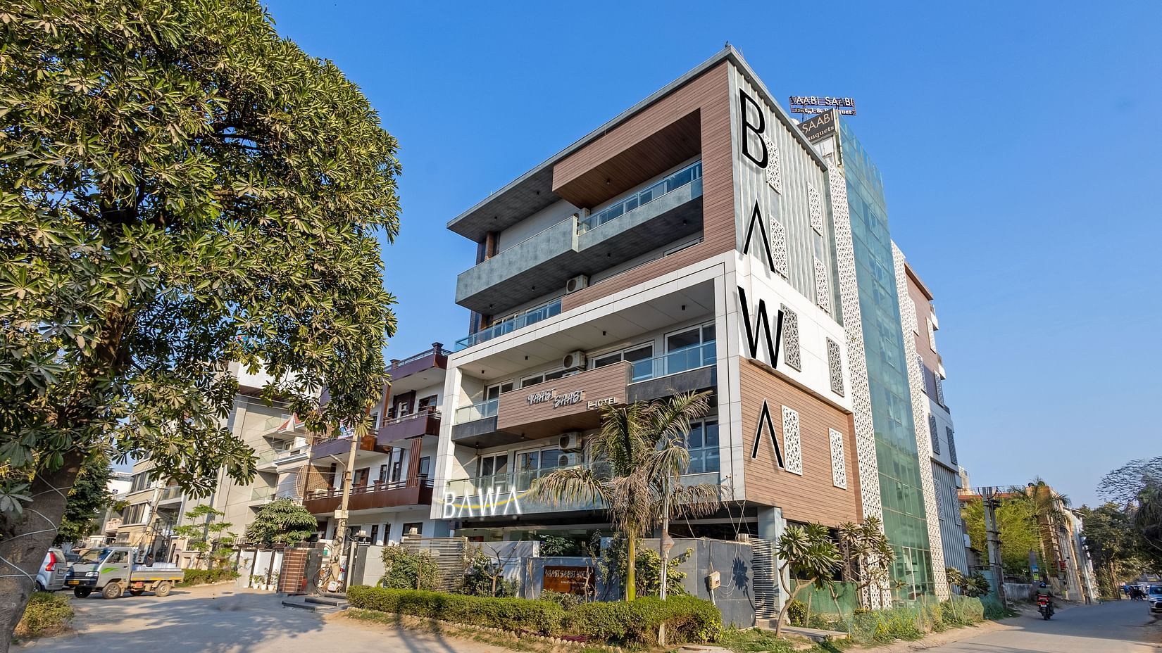 Exterior view of  the facade of Hotel Bawa, Gurgaon, a multi-storeyed building with a tree in the foreground and a clear blue sky in the background.