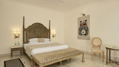 Bedroom of Superior Suites at Fateh Safari Suites, Kumbhalgarh featuring double size bed, a sofa, bedside table with lights on either side, a wooden chair, and a table.