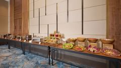 A long table laden with a variety of foods, from appetisers to desserts, is set up in a brightly lit room with patterned blue carpet, ready for guests to enjoy. - Iris Sarovar Portico, Sector 85, Gurugram