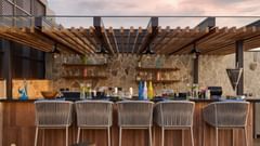 A well-stocked bar with a wooden counter and comfortable stools is prepared for service, with bottles and glassware neatly arranged on shelves beneath a slatted roof. - Iris Sarovar Portico, Sector 85, Gurugram