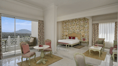 A bright and spacious Heritage Chamber Suite at Fateh Khas Mahal Suites, Udaipur features a sitting area with pink chairs and a small table, a bed in the background, and a large window offering a view.